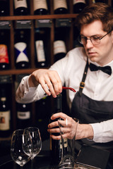 Bartender opening a wine bottle with corkscrew in restaurant with wine shelves.