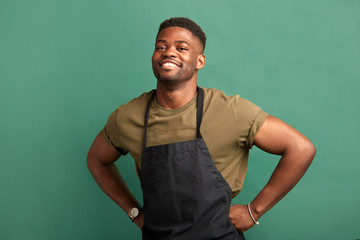 African male farmer with muscular hands and sportive body, dressed in apron smiling at camera with toothy smile over green background - obrazy, fototapety, plakaty