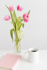 Bouquet of pink tulips in a glass vase, mug and books on a white table.