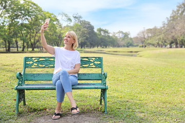 elderly caucasian woman wearing white shirt and blue jean using cell phone and self portrait in the park during summer time