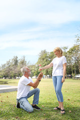 elderly caucasian couple with white shirt and blue jean sitting and standing in park during summer time while husband give red rose to wife