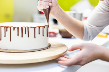 A confectioner squeezes liquid chocolate from a pastry bag onto a white cream biscuit cake on a wooden stand. The concept of homemade pastry, cooking cakes.