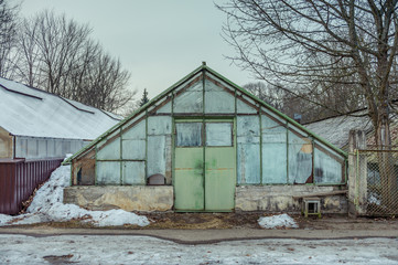 Old greenhouse made of glass with a triangular roof. Cloudy day in early spring.