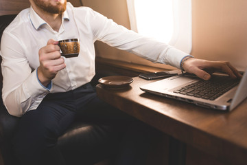 Attractive and successful businessman drinking coffee and working behind a laptop while sitting in a chair of his private jet.