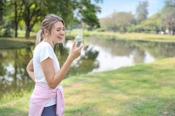 young beautiful caucasian woman wearing white sportswear standing in park. she is enjoying while drinking pure water with laughing and happy smiling face