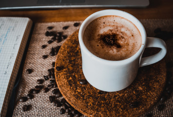 cup of coffee with foam with coffee bean, book, and laptop as a background
