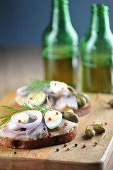Sandwiches with herring, onions, capers and quail eggs. Traditional dutch snack. In the background are two beer bottles. Dark background. Close-up. Vertical frame orientation.