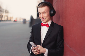 Naklejka premium Portrait of a young guy in a black suit and red bow-tie. man listening to music in big headphones.