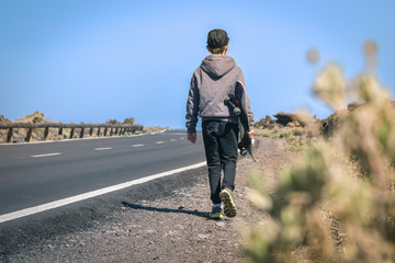 Obraz premium Trendy beautiful kid walk on the path side of the road with skateboard in hand. Back view of hipster trendy boy, walking holding skateboard enjoying journey. Concept of freedom and independence.
