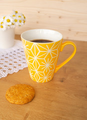 Fototapeta na wymiar View of a bright yellow cup of coffee, a cookie and a bunch or bouquet of blooming daisies in a white porcelain vase on wooden table against a wooden background. Hello spring or good morning coffee 