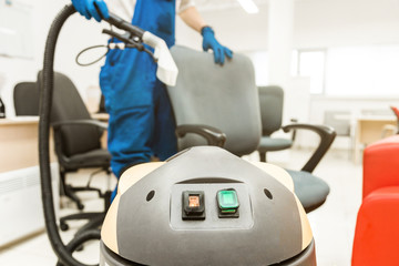 Young man in workwear and rubber gloves cleans the office chair with professional equipment.