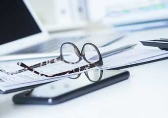 Closeup of white desktop with laptop, spectacles, notepads, pen, smartphone, clipboard.
