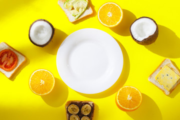 Obraz na płótnie Canvas Sandwiches with banana, tomato, avocado, cheese and fresh fruit, orange, coconut and an empty white plate on a yellow background. Concept of healthy breakfast, vitamins, diet. Flat lay, Top view.