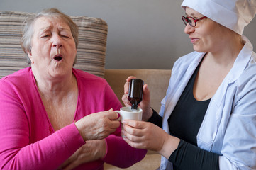 seriously ill elderly woman in pink blouse is sitting on sofa. Nurse girl in white medical gown...