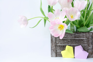 A bouquet of pink tulips in a wooden box and two paper hearts of yellow and lilac color on a white background.