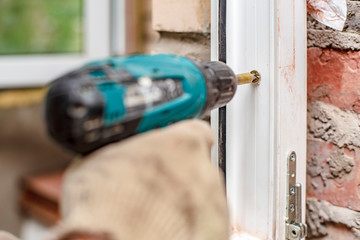 Male hands using an electric screwdriver and screws are going to fasten a PVC window frame to a...