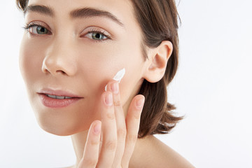 Obraz na płótnie Canvas Smiling young female applying cream on face skin