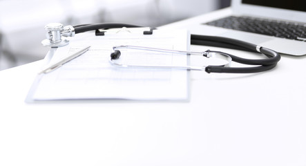 Stethoscope, clipboard with medical form lying on hospital reception desk with laptop computer and busy doctor and patient communicating at the background. Medical tools at doctor working table