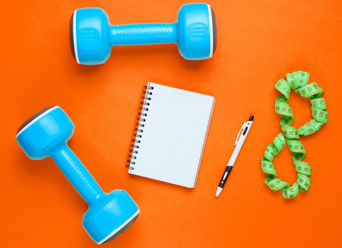 Fitness concept, workout plan. Plastic blue dumbbells, notepad, ruler on orange background. The idea of ​​losing weight. Top view. Flat lay