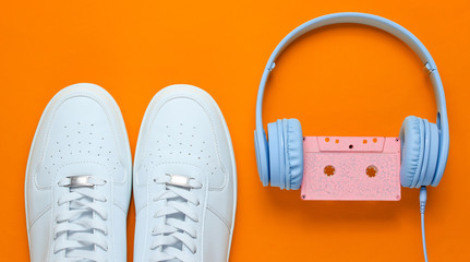 Retro style. 80s. Pop culture. Minimalismalism. Headphones with audio cassette, white sneakers on orange background. Top view