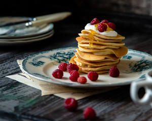 Homemade pancakes with honey, white cream, raspberries on vintage plate with green ornament on wooden rustic