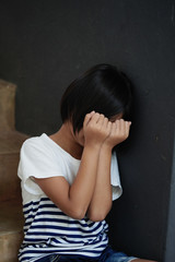 An Asian girl sat unhappy with her hands covering her face and leaning against the wall.