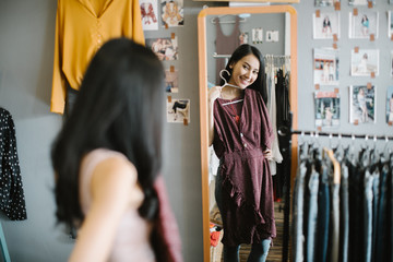 shopping, fashion, style and people concept - happy woman choosing clothes and looking to mirror in mall or clothing store