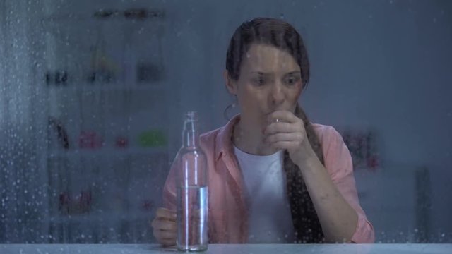 Sad woman with wounded face drinking vodka behind rainy window, addiction