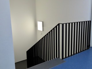 Detail of staircase in a modern building with white walls, blue floor, black railings, lit white square lamp on the wall