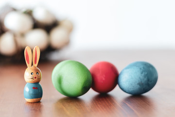 Small wooden easter rabbit beside three colorfull easter eggs on a wooden table 