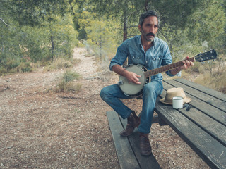 attractive man with beard playing on banjo in the forest sitting at a table - obrazy, fototapety, plakaty