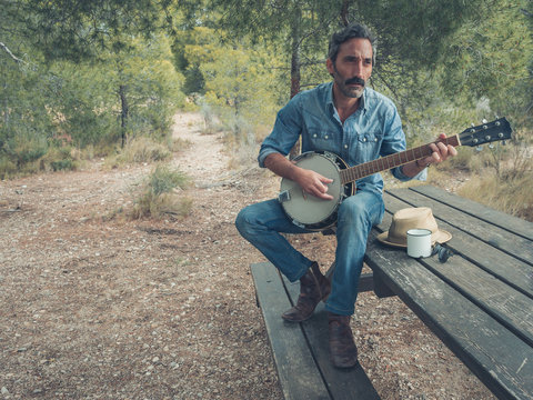 attractive man with beard playing on banjo in the forest sitting at a table