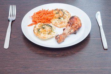 First person view for breakfast of fried scrambled eggs, chicken leg and Korean carrot on a white plate with a fork and knife on a wooden table.