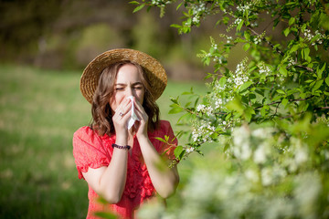 Young girl blowing nose and sneezing in tissue in front of blooming tree. Seasonal allergens affecting people. Beautiful lady has rhinitis.