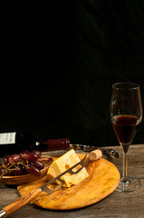 Top view, close distance of a round, rare wood plate with a wedge of french cheese, cheese knife and a wood bowl of red grapes  with a corkscrew and cork,  neck unopened bottle, red wine