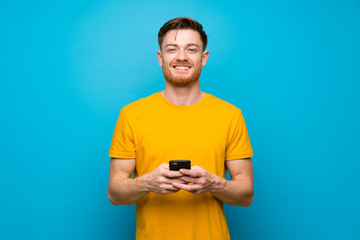 Redhead man over blue wall sending a message with the mobile