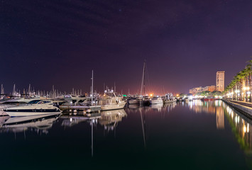 Fototapeta na wymiar Marine port in Alicante at night. Luxury yachts, ships and fishing boats are standing in rows in harbor. Costa Blanca, Spain, Mediterranean sea. Main focus on boats. Long exposure photo. Soft light