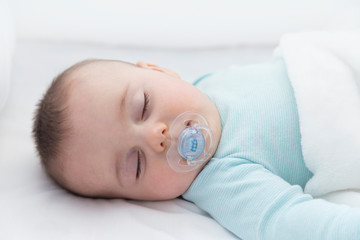 Baby sleeping with open arms in a cradle . Light blue pajama and white bed sheets. A pacifier in his mouth.