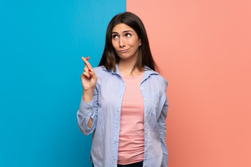 Young woman over pink and blue wall with fingers crossing and wishing the best