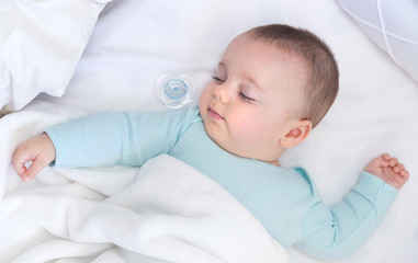 Baby sleeping with a pacifier aside. Light blue pajama and white bed sheets.