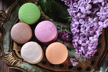 Multicolored bright macaroons on a wooden vintage tray next to flowers of lilac on a wooden table, top view