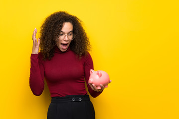 Dominican woman with turtleneck sweater surprised while holding a piggybank - obrazy, fototapety, plakaty