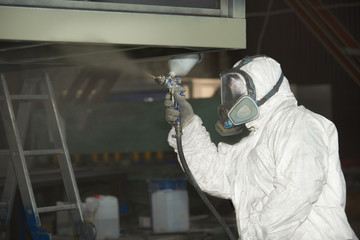 A man in a white uniform applies paint with a spray gun on a metal product.