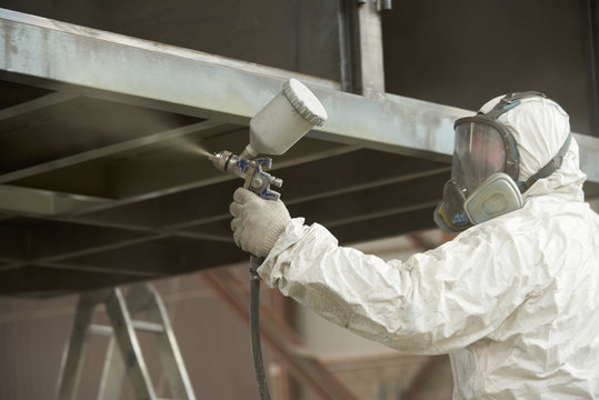 A man in a white uniform applies paint with a spray gun on a metal product.