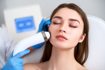 Beautician doctor doing rf-lifting procedure for flawless woman face laying in a beauty salon....