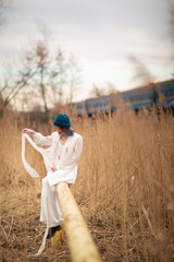 A young girl dressed in a white long dress, sitting on a pipe, near a wheat field. Behind her is a train