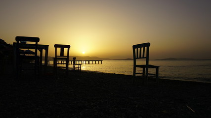 Empty wooden chairs on pebble beach during dramatic sunset. Summer is coming and outdoor / outside conceptual photo. Amazing sunset over mountains & ocean horizon at weekend. Sunny day at outdoor cafe