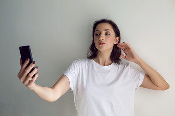 girl in a white t-shirt takes a selfie on a black phone against the white wall