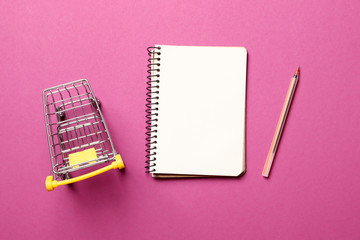 Shopping concept. shopping cart, blank paper notebook with pen on a pink background.