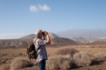 Side view of the senior man with backpack looking with a binoculars the landscape around. Volcanic...
